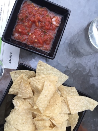 A black square dish contains vibrant red salsa with visible herbs and small chunks of vegetables, positioned next to a black rectangular dish filled with a pile of yellow tortilla chips. Both dishes rest on a glass table, and a glass of water is partially visible on the side.