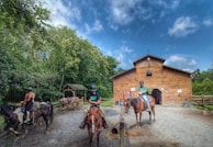 A wooden barn is situated in a rural setting with a dirt area in front, where two individuals on horseback are positioned. The area is surrounded by lush green trees under a clear blue sky with scattered clouds. A horse tied under a wooden shelter can be seen in the background.