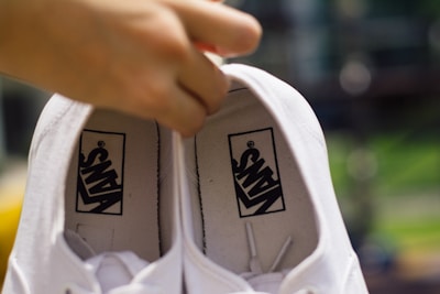 A pair of white shoes are being held by a hand from inside the shoe. The shoes have black logos printed inside with the brand name. The background appears blurred with hints of green and yellow colors.