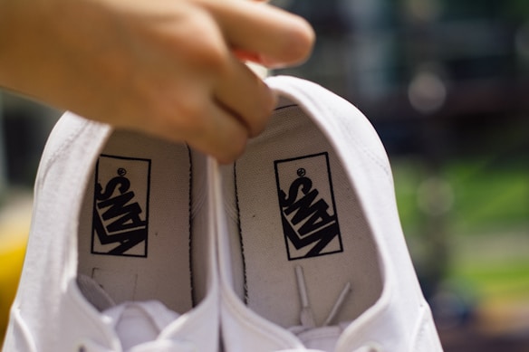 A pair of white shoes are being held by a hand from inside the shoe. The shoes have black logos printed inside with the brand name. The background appears blurred with hints of green and yellow colors.