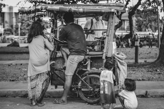 A group of people gathered around a makeshift tricycle or pedicab, which appears to be parked in a park or outdoor area. The scene includes an adult man, a woman, and children, suggesting a family or community setting. Trees and a building are visible in the background, contributing to an urban outdoor environment.