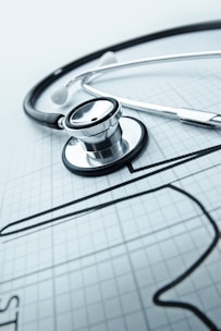 A stethoscope is placed on a sheet of graph paper, which features a sketched outline of a heart. The stethoscope lies diagonally across the paper, and its metallic and rubber components are clearly visible, suggesting a medical or healthcare theme.