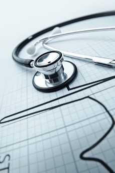 A stethoscope is placed on a sheet of graph paper, which features a sketched outline of a heart. The stethoscope lies diagonally across the paper, and its metallic and rubber components are clearly visible, suggesting a medical or healthcare theme.