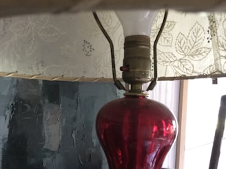 A vintage-style lamp with a red glass base and a light bulb beneath a patterned fabric lampshade. The lampshade features a floral design with leaves and small flowers. The background includes a textured, dark wall, possibly with an abstract painting or uneven finish.
