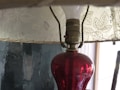 A vintage-style lamp with a red glass base and a light bulb beneath a patterned fabric lampshade. The lampshade features a floral design with leaves and small flowers. The background includes a textured, dark wall, possibly with an abstract painting or uneven finish.
