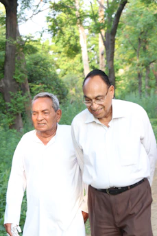 Two elderly men walking together on a path surrounded by lush green trees and foliage. Both appear to be enjoying a conversation and are dressed in light-colored clothing.