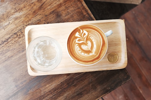 A wooden tray sitting on a rustic wooden table holds a cup of latte with intricate latte art. Next to the latte is a glass of water, and a gold-colored coin or token.