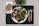 A plate of mixed green salad with grilled vegetables on a black placemat, accompanied by a whole grain bread roll on a napkin. Wooden fork and knife are placed to the side, with a small green plant in the background.