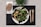 A plate of mixed green salad with grilled vegetables on a black placemat, accompanied by a whole grain bread roll on a napkin. Wooden fork and knife are placed to the side, with a small green plant in the background.