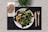 A plate of mixed green salad with grilled vegetables on a black placemat, accompanied by a whole grain bread roll on a napkin. Wooden fork and knife are placed to the side, with a small green plant in the background.