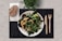 A plate of mixed green salad with grilled vegetables on a black placemat, accompanied by a whole grain bread roll on a napkin. Wooden fork and knife are placed to the side, with a small green plant in the background.
