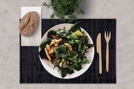 A plate of mixed green salad with grilled vegetables on a black placemat, accompanied by a whole grain bread roll on a napkin. Wooden fork and knife are placed to the side, with a small green plant in the background.