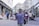 A street musician with a guitar is performing for a diverse group of pedestrians on a busy city street. The scene includes people walking, others stopping to listen or watch the performance, and various storefronts lining the sides of the street. The atmosphere is lively and filled with movement, capturing a moment of everyday urban life.