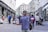 A street musician with a guitar is performing for a diverse group of pedestrians on a busy city street. The scene includes people walking, others stopping to listen or watch the performance, and various storefronts lining the sides of the street. The atmosphere is lively and filled with movement, capturing a moment of everyday urban life.