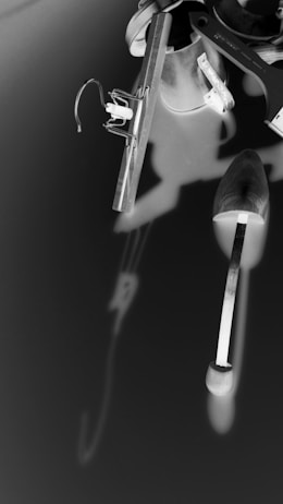 A monochrome image featuring a collection of tools and objects, including a mixing spatula, a saw, and a clip attached to a wire. The objects appear to be artistically arranged and create interesting shadows against the contrasting background.