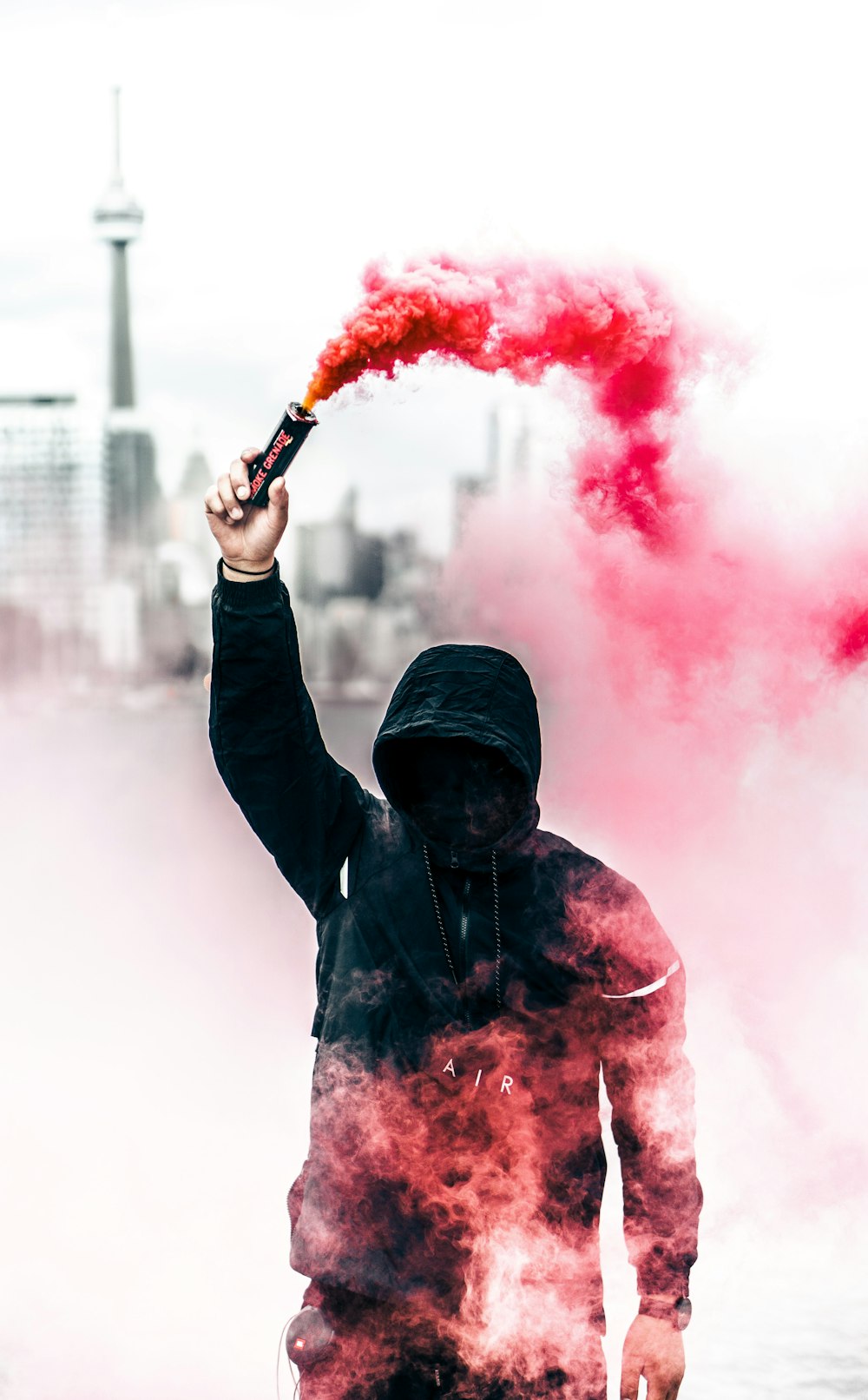 A person in a black hoodie with obscured face holds up a pink smoke grenade