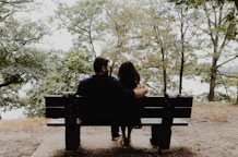 man looking to woman sitting on black wooden bench in front of tall trees during daytime