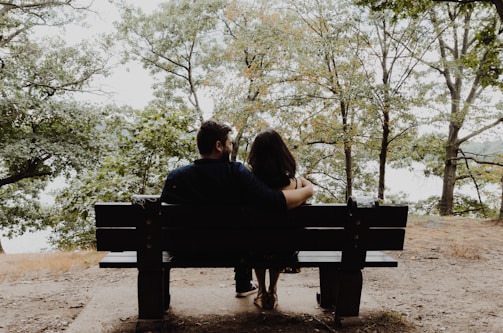 man looking to woman sitting on black wooden bench in front of tall trees during daytime