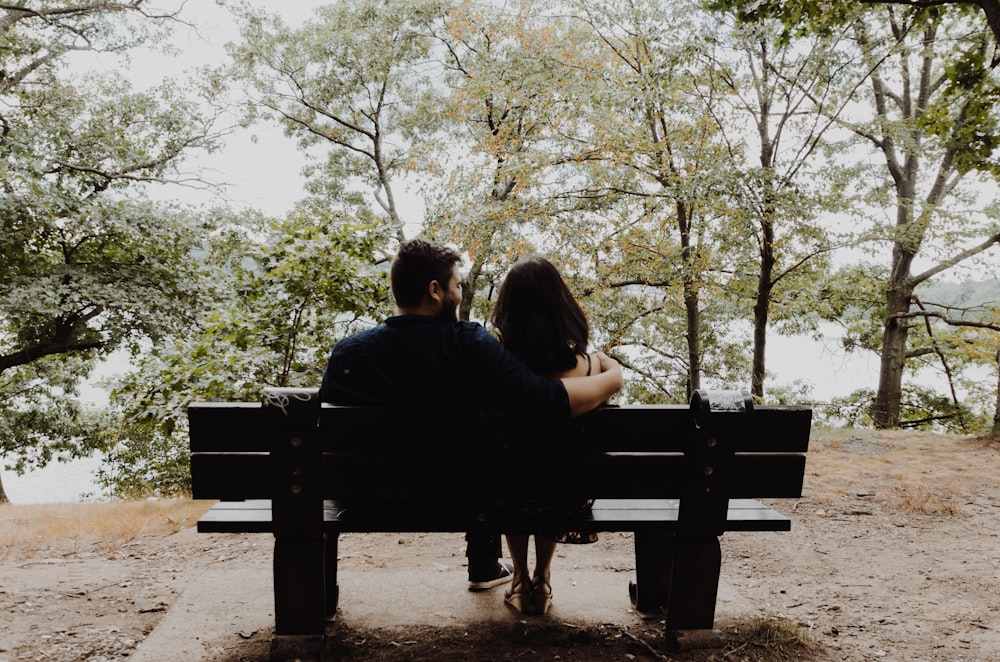 man looking to woman sitting on black wooden bench in front of tall trees during daytime