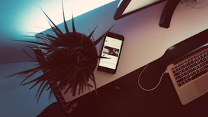 A modern workspace features a white desk with a potted plant, a smartphone displaying content, and an open laptop. The lighting creates a contrasting shadow effect, lending a dramatic ambiance. The scene combines technology and a hint of nature.