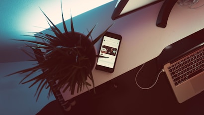 A modern workspace features a white desk with a potted plant, a smartphone displaying content, and an open laptop. The lighting creates a contrasting shadow effect, lending a dramatic ambiance. The scene combines technology and a hint of nature.