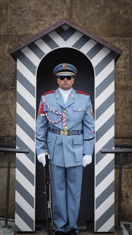 A uniformed guard stands in a small sentry box adorned with a striped pattern. The guard is wearing a formal blue-gray uniform with decorative cords and insignia, complete with a peaked cap and sunglasses. He is holding a rifle, and the sentry box is positioned against a stone wall.