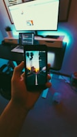 A hand holds a smartphone displaying a lock screen with a scenic image of palm trees and a sunset. The screen shows the time '1:37' and date 'Saturday, August 12'. In the background, there is a modern workspace setup with a computer monitor, laptop, and other office items on a desk. The room is dimly lit with a cool, bluish backlight.