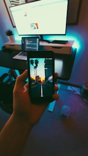 A hand holds a smartphone displaying a lock screen with a scenic image of palm trees and a sunset. The screen shows the time '1:37' and date 'Saturday, August 12'. In the background, there is a modern workspace setup with a computer monitor, laptop, and other office items on a desk. The room is dimly lit with a cool, bluish backlight.