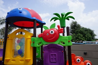 A colorful playground structure features whimsical designs with a variety of shapes and colors, including a red apple with a face, a green leaf, and a small plastic palm tree. The playset includes a yellow window with gears on the side and is topped with a red and blue dome resembling a ladybug. The background shows a cloudy sky and some foliage.