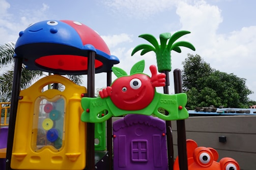 A colorful playground structure features whimsical designs with a variety of shapes and colors, including a red apple with a face, a green leaf, and a small plastic palm tree. The playset includes a yellow window with gears on the side and is topped with a red and blue dome resembling a ladybug. The background shows a cloudy sky and some foliage.