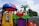 A colorful playground structure features whimsical designs with a variety of shapes and colors, including a red apple with a face, a green leaf, and a small plastic palm tree. The playset includes a yellow window with gears on the side and is topped with a red and blue dome resembling a ladybug. The background shows a cloudy sky and some foliage.