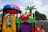A colorful playground structure features whimsical designs with a variety of shapes and colors, including a red apple with a face, a green leaf, and a small plastic palm tree. The playset includes a yellow window with gears on the side and is topped with a red and blue dome resembling a ladybug. The background shows a cloudy sky and some foliage.