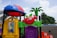 A colorful playground structure features whimsical designs with a variety of shapes and colors, including a red apple with a face, a green leaf, and a small plastic palm tree. The playset includes a yellow window with gears on the side and is topped with a red and blue dome resembling a ladybug. The background shows a cloudy sky and some foliage.