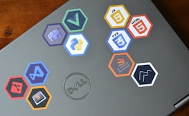 A laptop is adorned with various hexagonal stickers featuring different programming languages and tools, such as PHP, Python, JavaScript, HTML, CSS, Git, and others. The central logo on the laptop is from Dell.