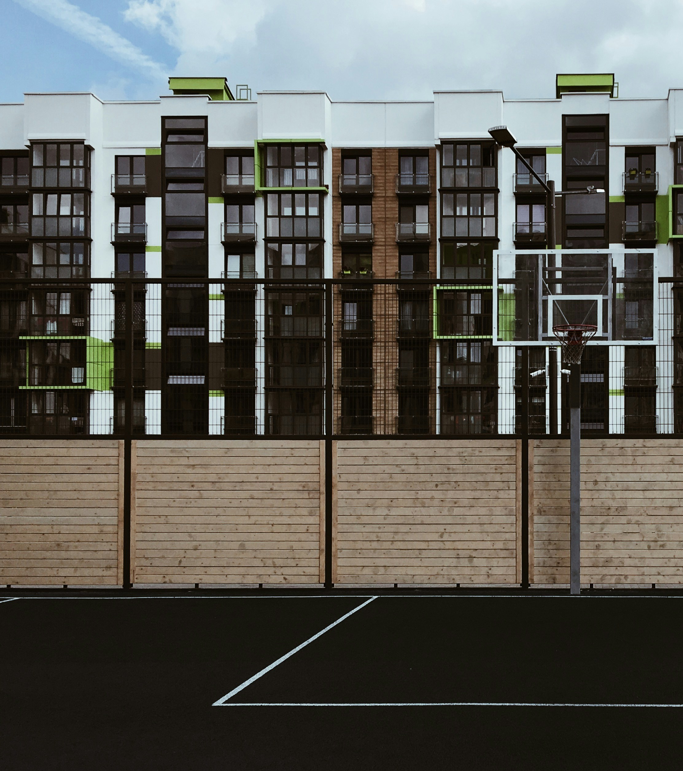 En primer plano se ve una cancha de baloncesto con red, caracterizada por una superficie negra con líneas blancas. Detrás hay una valla de madera y, más allá, un moderno edificio de apartamentos con una mezcla de detalles en negro, blanco y verde. El cielo está parcialmente nublado.