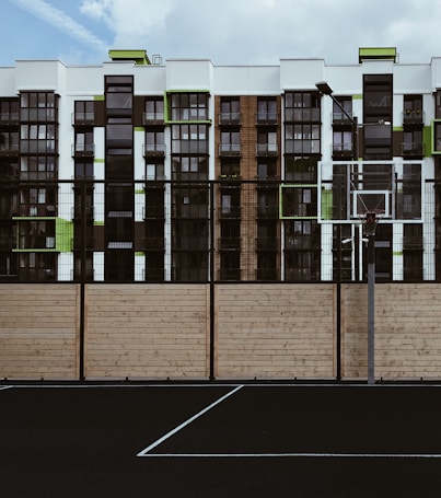 A basketball court with a net is in the foreground, characterized by a black surface with white lines. Behind it, there is a wooden fence, and beyond that, a modern apartment building featuring a mix of black, white, and green accents. The sky is partly cloudy.