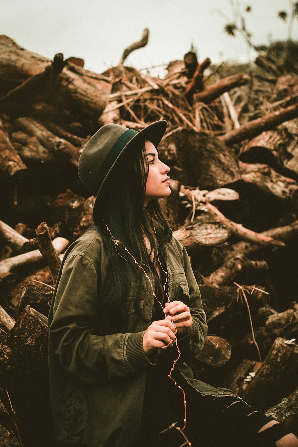mulher vestindo jaqueta bomber e chapéu lateral vendo na frente de troncos de madeira