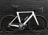 A sleek, modern road bicycle with a white frame and black handlebars, positioned against a dark brick wall. The bike features large wheels with the brand 'Shimano' visible on the crankset and a water bottle attached to the frame.