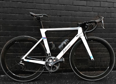 A sleek, modern road bicycle with a white frame and black handlebars, positioned against a dark brick wall. The bike features large wheels with the brand 'Shimano' visible on the crankset and a water bottle attached to the frame.