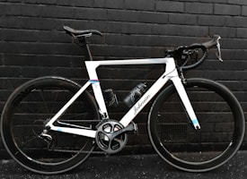 A sleek, modern road bicycle with a white frame and black handlebars, positioned against a dark brick wall. The bike features large wheels with the brand 'Shimano' visible on the crankset and a water bottle attached to the frame.