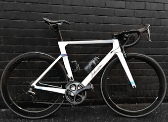 A sleek, modern road bicycle with a white frame and black handlebars, positioned against a dark brick wall. The bike features large wheels with the brand 'Shimano' visible on the crankset and a water bottle attached to the frame.