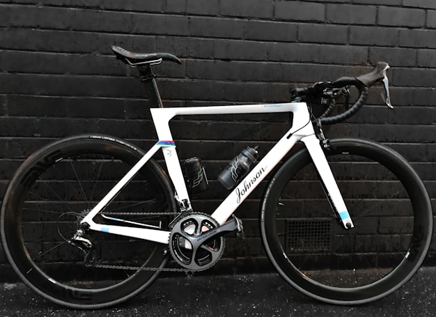A sleek, modern road bicycle with a white frame and black handlebars, positioned against a dark brick wall. The bike features large wheels with the brand 'Shimano' visible on the crankset and a water bottle attached to the frame.