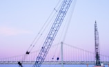 A large blue crane is positioned in front of a suspension bridge with a gentle evening sky in the background. Another crane or construction structure is visible at the right edge, indicating ongoing work or development. The scene captures elements of industrial activity against a serene backdrop.