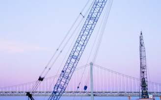 A large blue crane is positioned in front of a suspension bridge with a gentle evening sky in the background. Another crane or construction structure is visible at the right edge, indicating ongoing work or development. The scene captures elements of industrial activity against a serene backdrop.