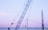 A large blue crane is positioned in front of a suspension bridge with a gentle evening sky in the background. Another crane or construction structure is visible at the right edge, indicating ongoing work or development. The scene captures elements of industrial activity against a serene backdrop.