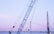 A large blue crane is positioned in front of a suspension bridge with a gentle evening sky in the background. Another crane or construction structure is visible at the right edge, indicating ongoing work or development. The scene captures elements of industrial activity against a serene backdrop.