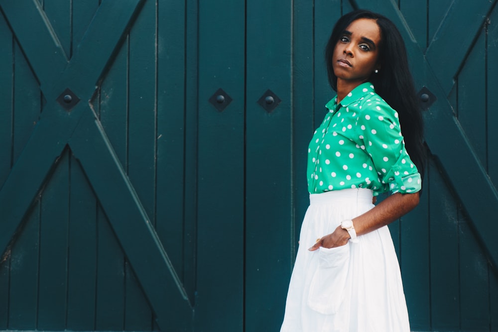 woman in white skirt and green dress shirt hand on pocket standing at the front of green door