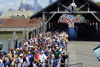 A large crowd of people is waiting in line along a wooden boardwalk next to a body of water. They are diverse in age and attire, with some wearing hats and sunglasses, hinting at a warm day. The line extends under a covered pathway, leading towards a distant urban skyline that includes towering skyscrapers. The atmosphere appears busy and bustling, typical of a popular tourist attraction.