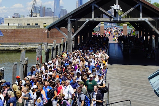 A large crowd of people is waiting in line along a wooden boardwalk next to a body of water. They are diverse in age and attire, with some wearing hats and sunglasses, hinting at a warm day. The line extends under a covered pathway, leading towards a distant urban skyline that includes towering skyscrapers. The atmosphere appears busy and bustling, typical of a popular tourist attraction.
