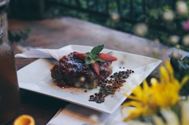 A beautifully plated dessert featuring a slice of cake topped with glossy red sauce, garnished with fresh strawberries and mint leaves. The cake rests on a white square plate placed on a wooden table, with a napkin to the side. There are chocolate sprinkles and crumbs scattered artistically around the slice. In the foreground, the image is flanked by blurred flowers, adding a touch of nature.